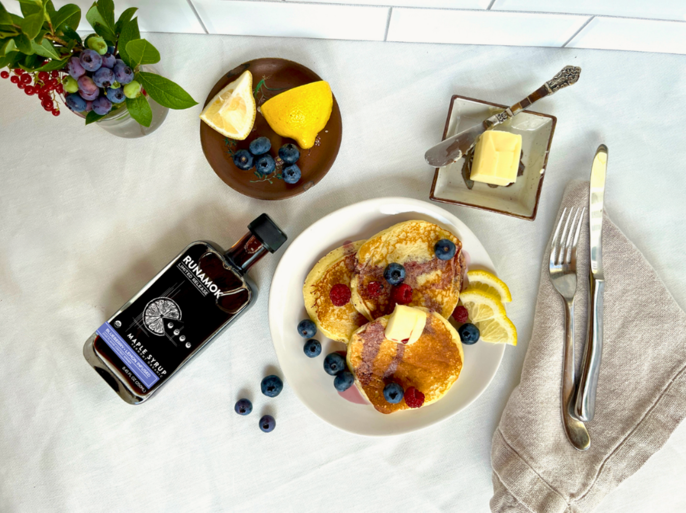 Breakfast setting with pancakes, berries, and a bottle of Runamok Maple Syrup on a white surface.