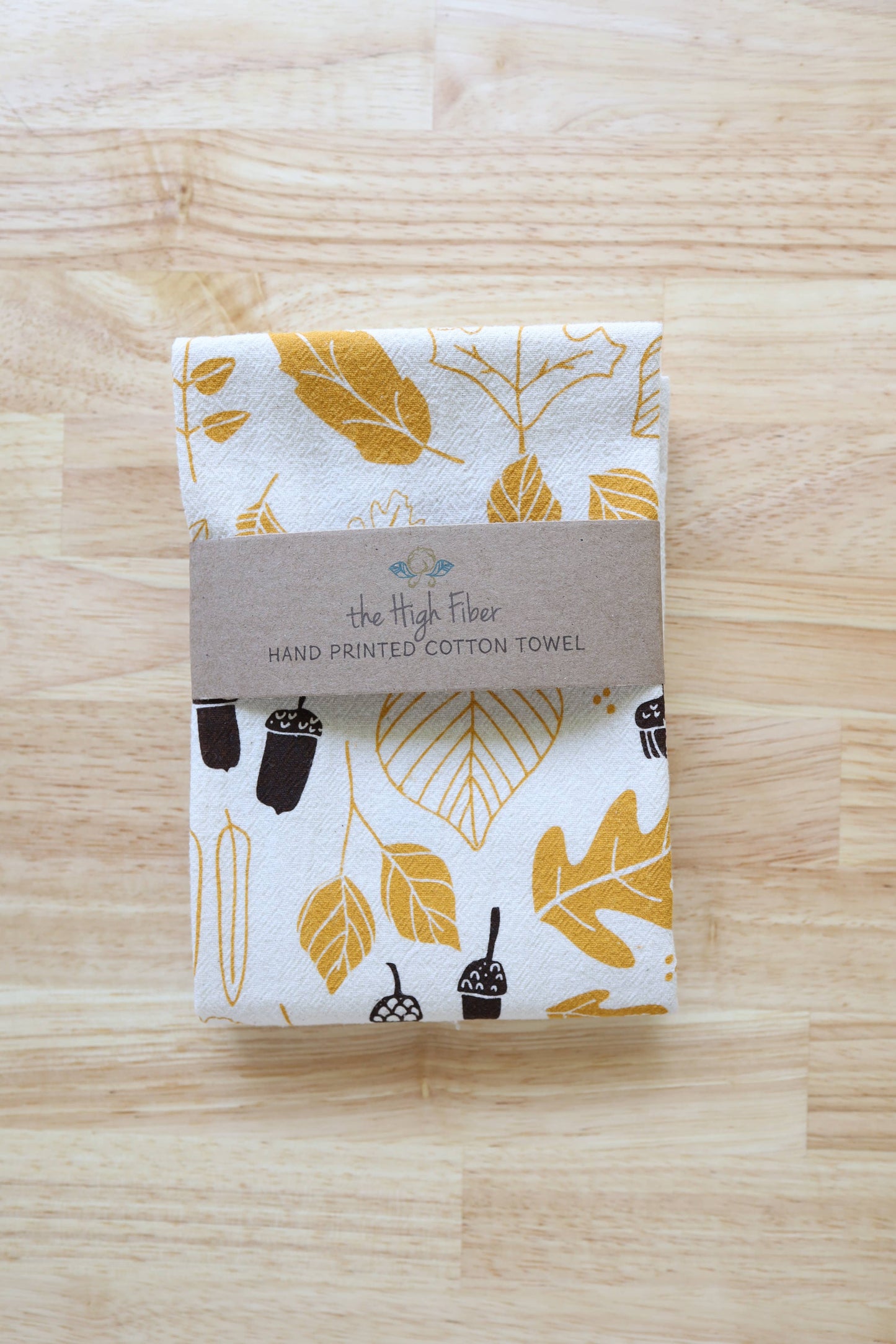 A folded cotton towel with a mustard yellow and dark brown botanical leaf print, wrapped in a brown paper band labeled 'the High Fiber', placed on a light wood background.