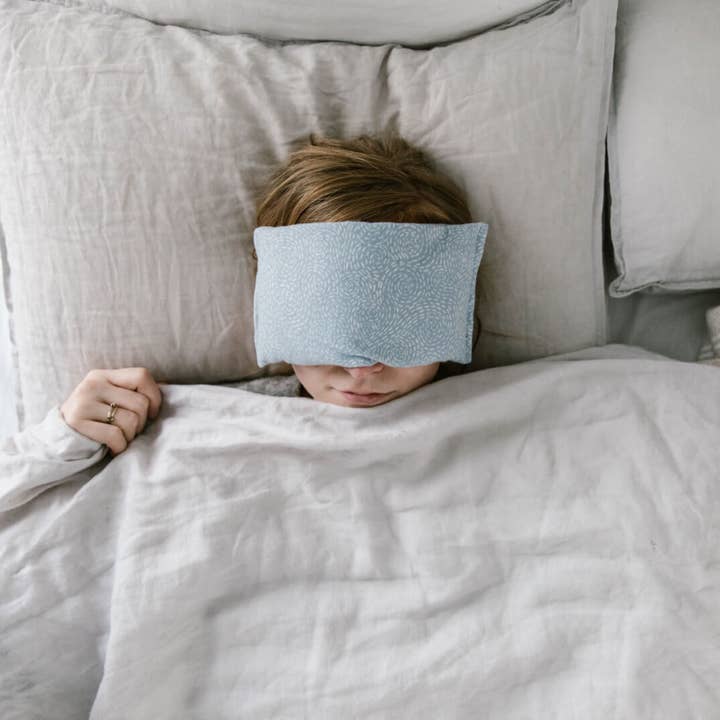 Person lying in bed with an eye pillow on on, partially covered by a blanket.