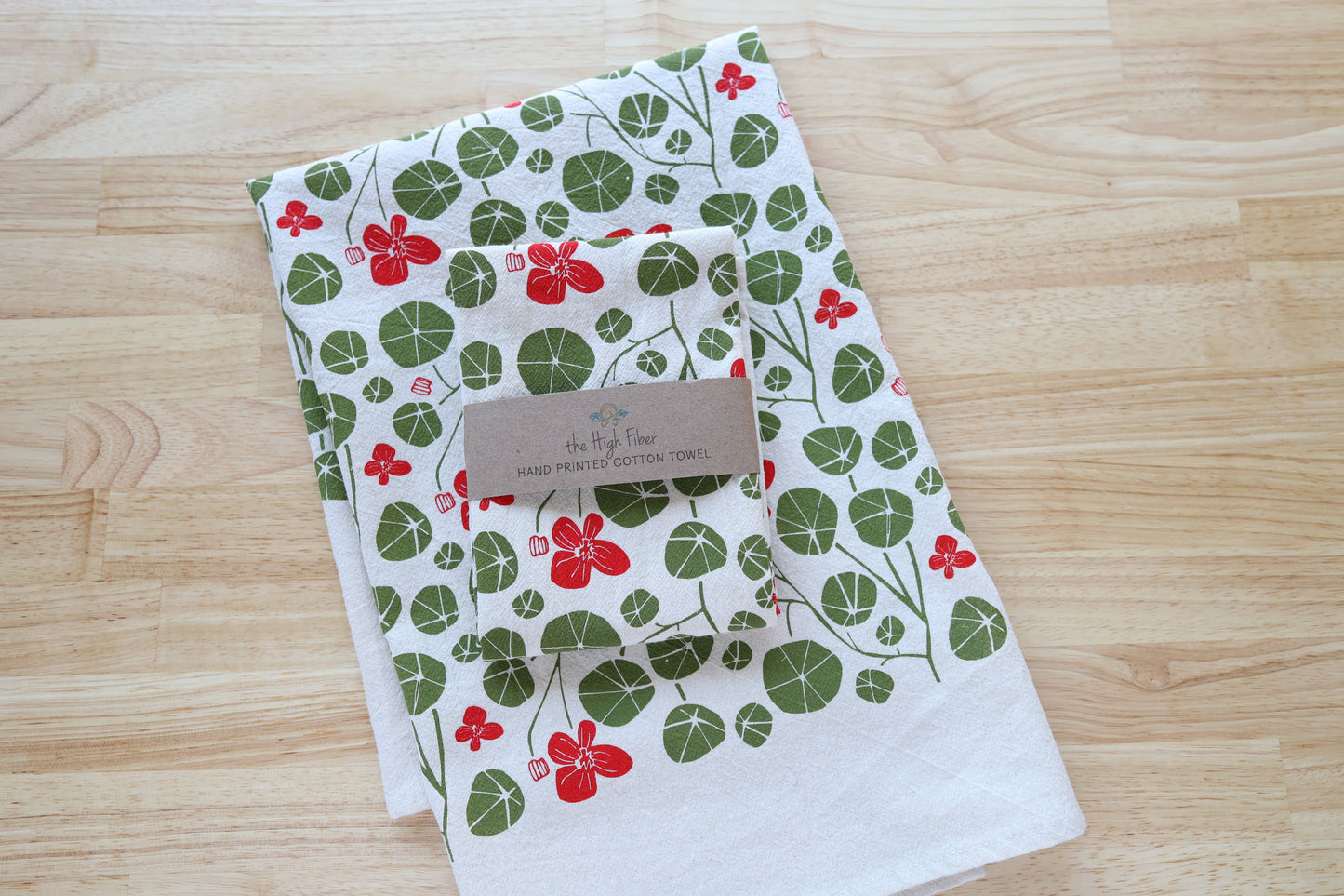 A folded white cotton tea towel with a green and red botanical pattern of flowers and leaves, resting on a light wood surface. A small paper tag is attached to the center.