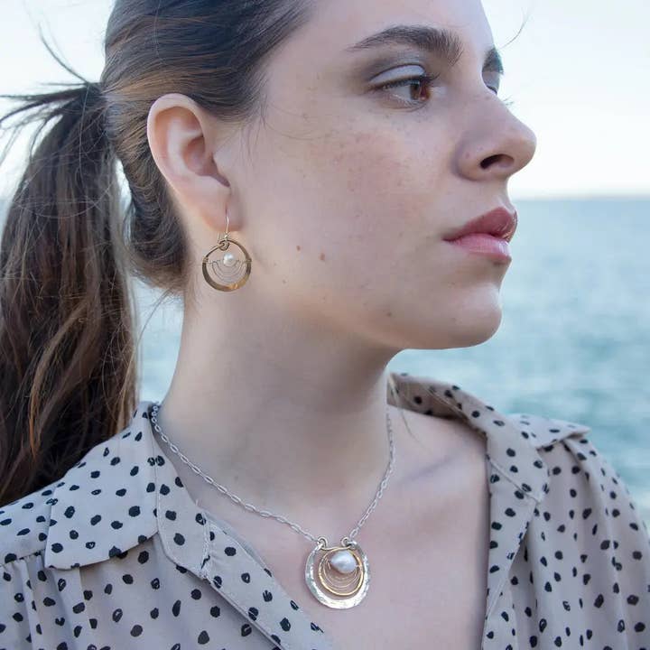 Woman wearing gold hoop earrings and a necklace with a pendant, standing by the ocean.