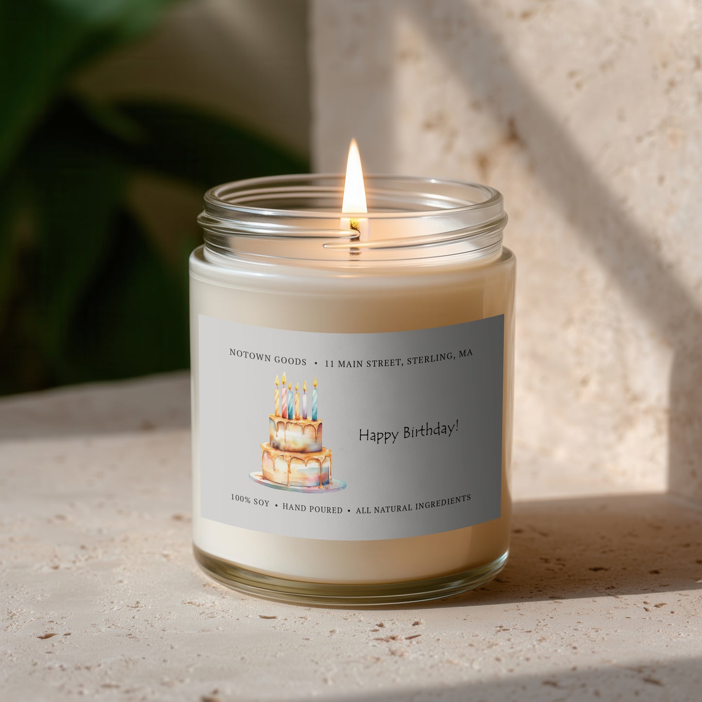Clear jar candle with white wax and a decorative label on a blurred outdoor background.