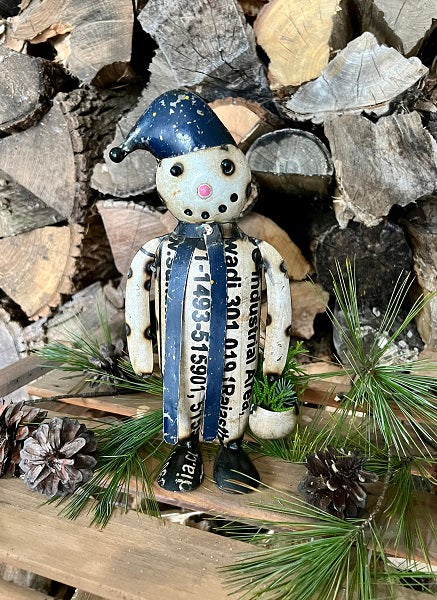 Decorative snowman figure with a blue hat and scarf on a wooden shelf with pinecones and greenery.
