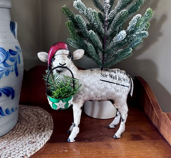 Decorative sheep figurine with a Santa hat and green basket, standing on a wooden surface next to a blue and white vase and a small Christmas tree.