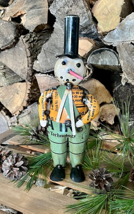 Decorative snowman figurine with a top hat standing on a wooden platform with pine cones and branches.