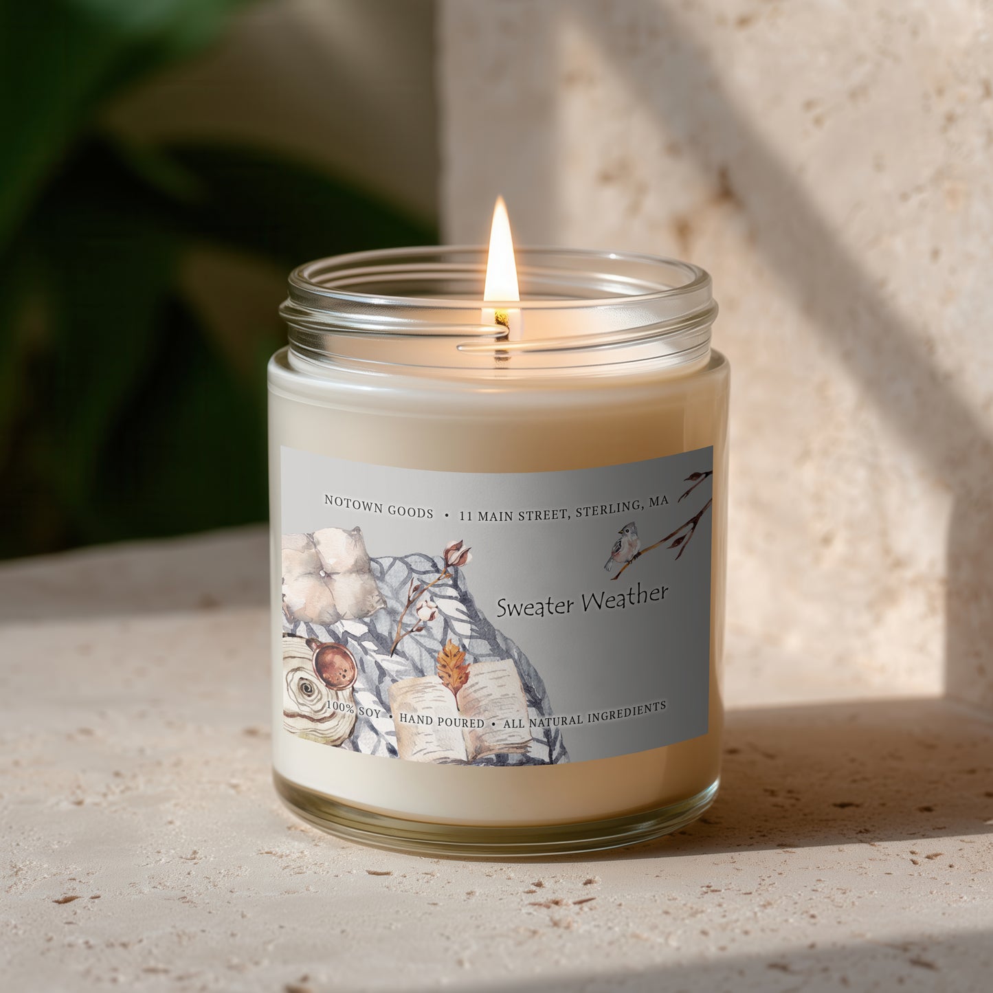 Clear jar candle with white wax and a decorative label on a blurred outdoor background.