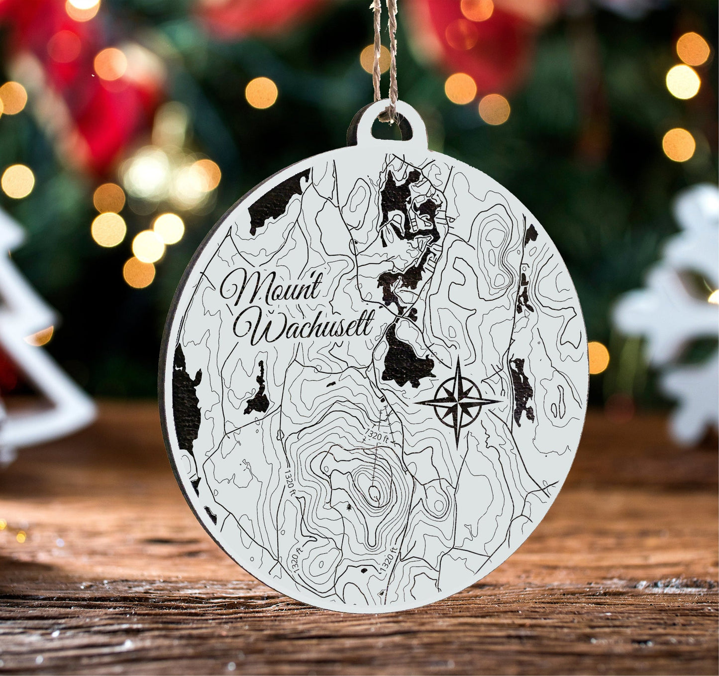 Decorative ornament with topographic design and 'Mount Wachusett' text on a wooden surface with Christmas lights in the background.