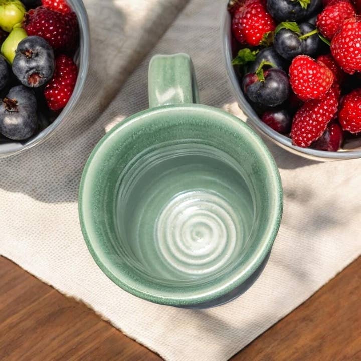 Green ceramic mug on a wooden surface with bowls of berries in the background