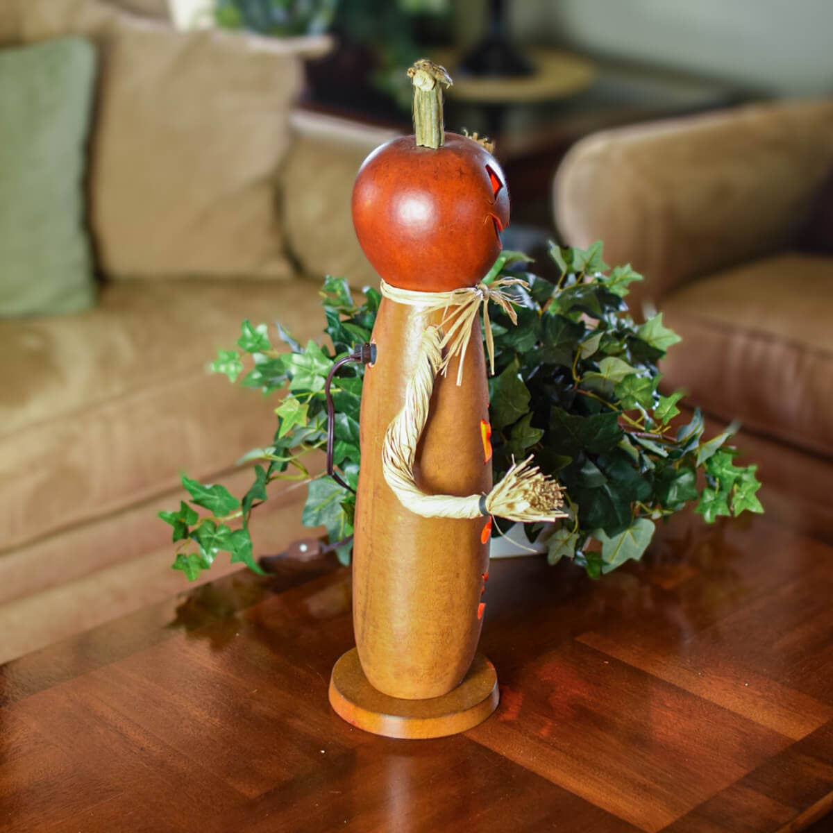 A decorative wooden scarecrow figure with a gourd head and straw details, standing on a wooden table in a living room setting with a sofa and greenery in the background.