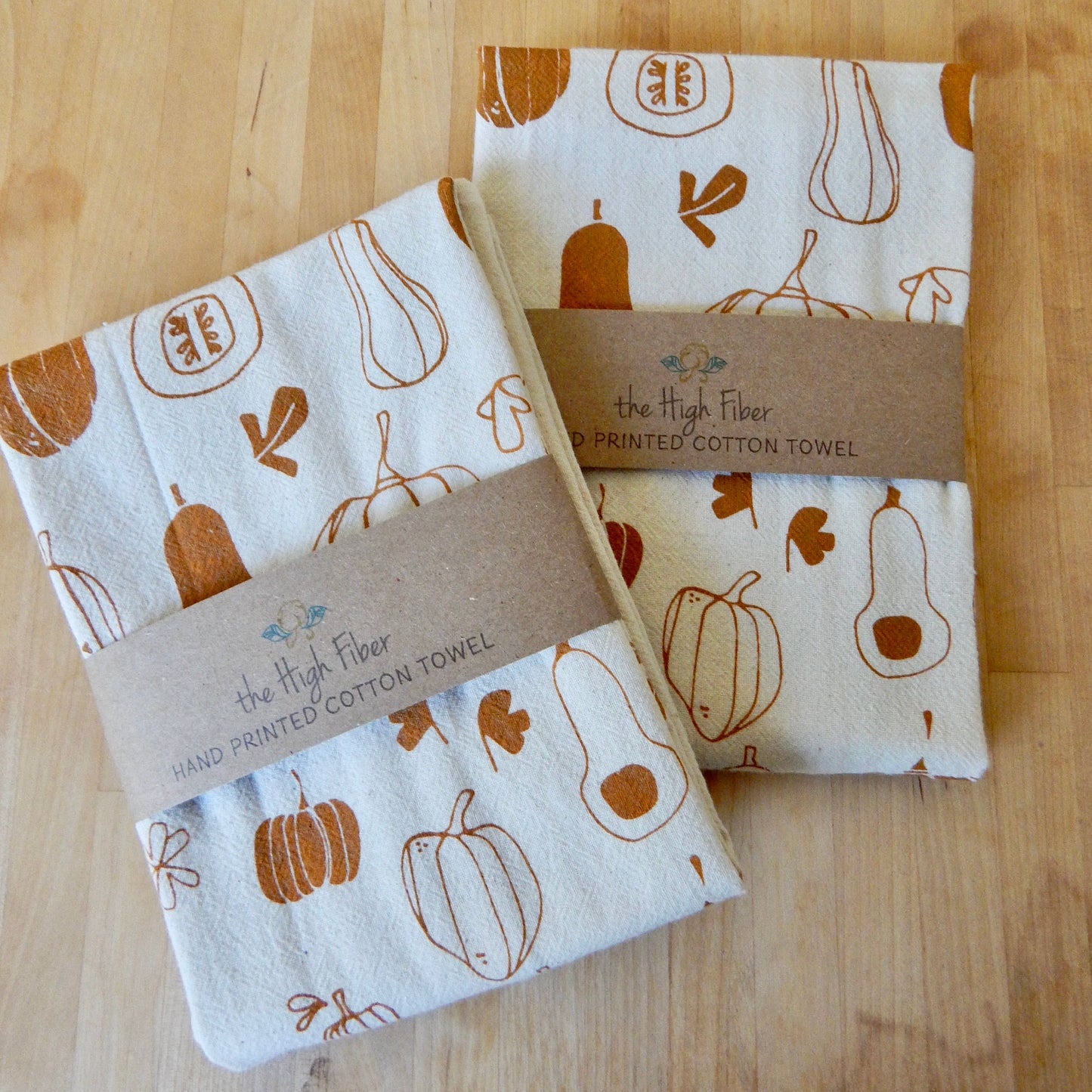Two natural cotton kitchen towels with a handprinted winter squash pattern, displayed on a wooden surface.