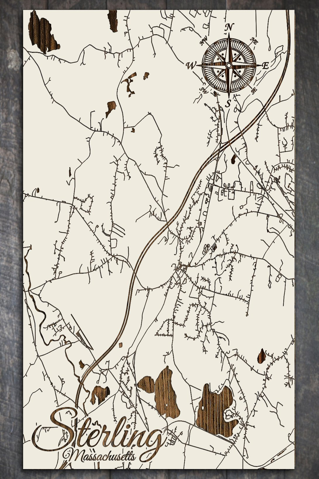 A wooden map of Sterling, MA, with cream-colored background and dark lines representing roads and landmarks, featuring a compass rose and the name 'Sterling, Massachusetts' at the bottom.