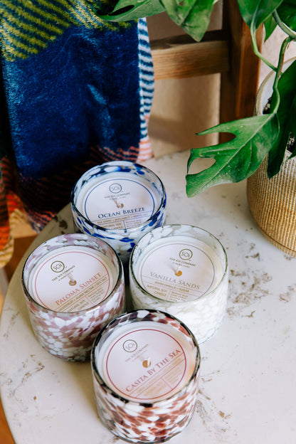 Five scented candles with decorative labels on a white surface, with a blanket and plant in the background.