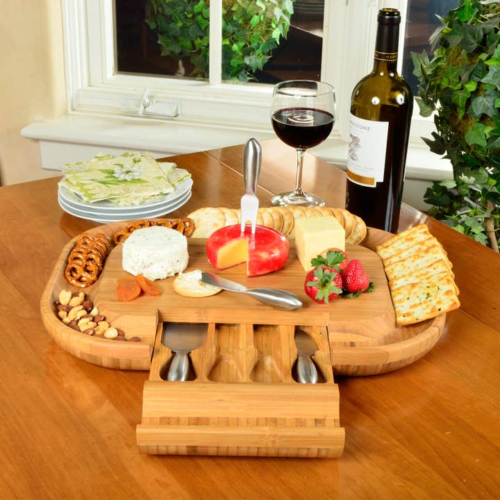 Wooden cheese board set with fruits, nuts, and a bottle of wine on a table.