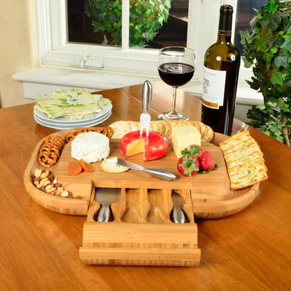 Wooden cheese board set with fruits, nuts, and a bottle of wine on a table.