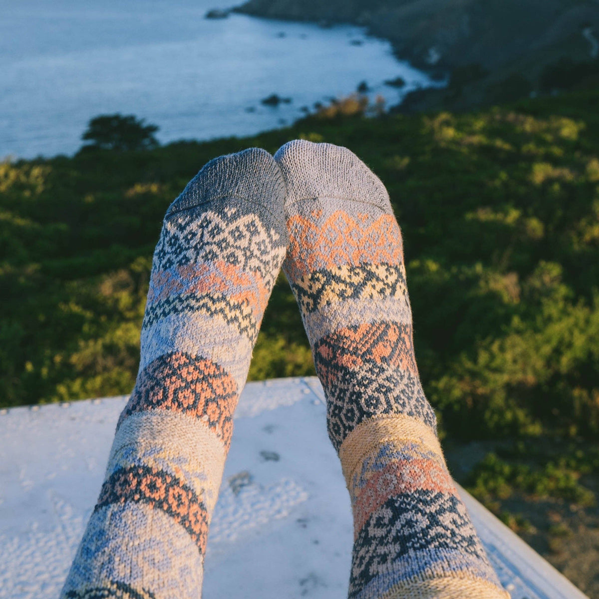 A pair of patterned knit socks worn on feet, photographed from above with an outdoor coastal landscape in the background.