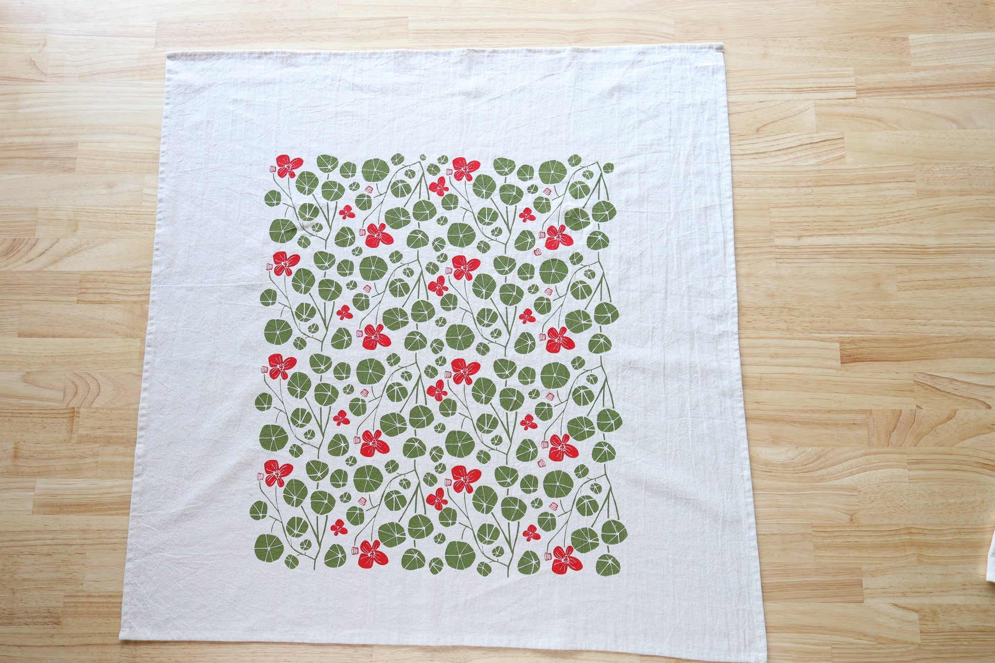 A white kitchen towel laid flat on a light wood floor, featuring a central rectangular print of green clover-like leaves and small red flowers.