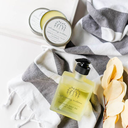 Natural body oil bottle and hand and cuticle salve tins with a gray and white striped towel and yellow flowers on a white background