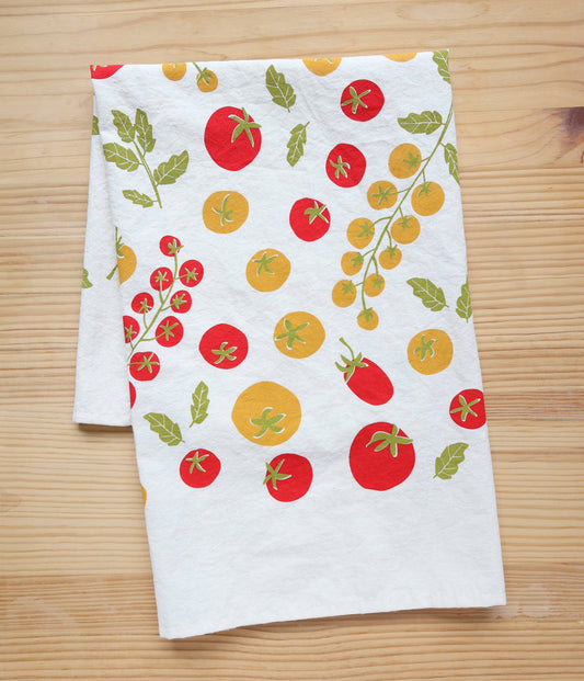 A white kitchen tea towel with a colorful tomato print, laid flat on a light wood grain surface.