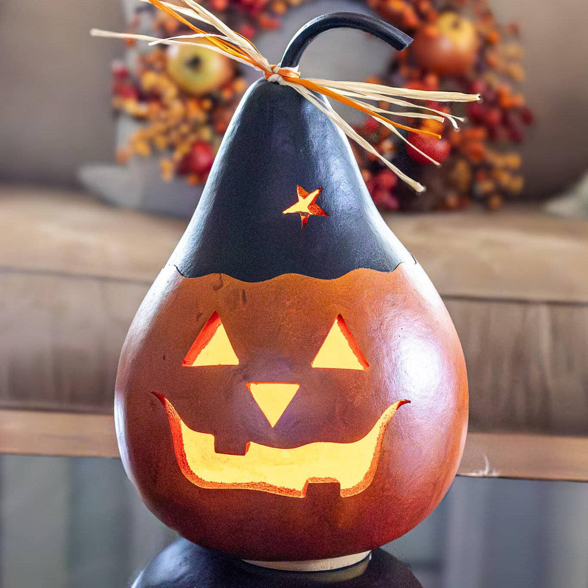A carved gourd pumpkin with a glowing jack-o'-lantern face, topped with a black pointed hat and orange straw accents, photographed in a softly blurred indoor setting.