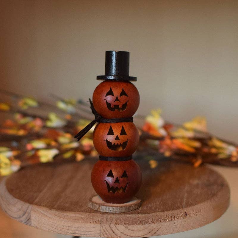 A small Halloween decoration made of three stacked orange jack-o'-lantern gourds with carved faces, a black top hat, and a black ribbon bow, displayed on a round wooden stand with blurred autumn leaves in the background.