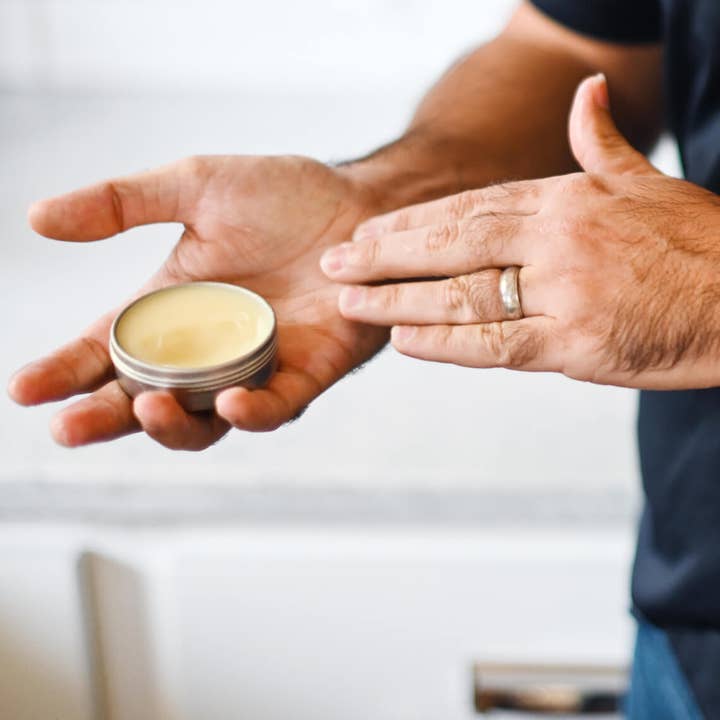 Person holding a small container of hand and cuticle salve with a blurred background