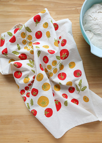 A white kitchen tea towel with a colorful print of red and yellow tomatoes and green leaves, draped on a light wood table next to a bowl of flour.