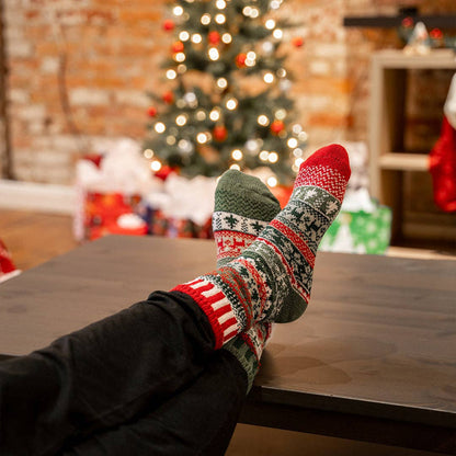 A person's feet wearing a pair of green, red, and white patterned knit Christmas socks resting on a dark wooden table, with a blurred Christmas tree in the background.