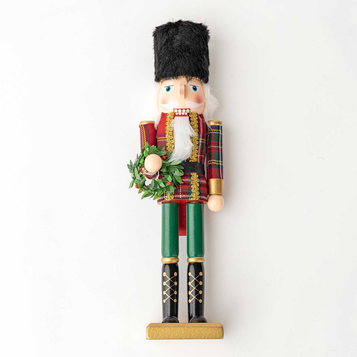 A traditional wooden nutcracker figurine standing on a gold-colored base, wearing a red and green plaid coat, green trousers, black boots, and a black fur hat, holding a green wreath.