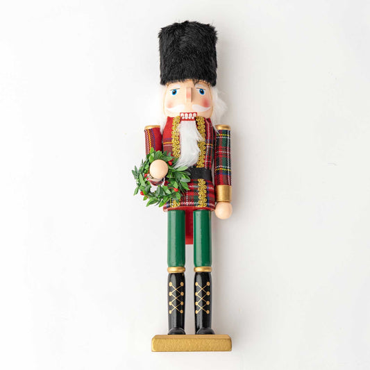 A traditional wooden nutcracker figurine standing on a gold-colored base, wearing a red and green plaid coat, green trousers, black boots, and a black fur hat, holding a green wreath.