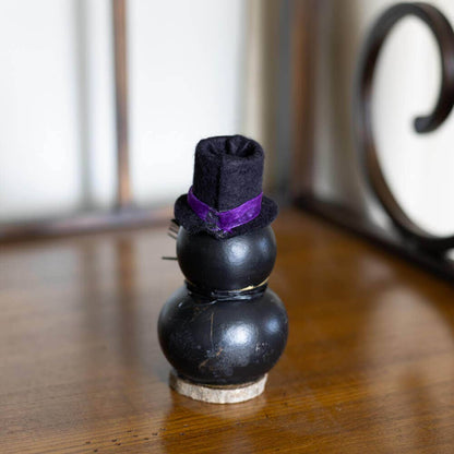 A small black gourd figurine wearing a black felt top hat with a purple band, standing on a wooden surface with a blurred indoor background.