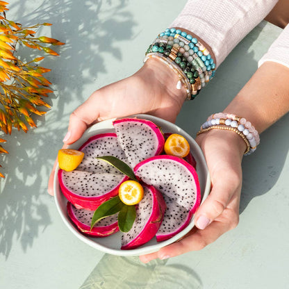 Person holding a bowl of dragon fruit slices with lemons on a light background, wearing colorful bracelets on both wrists.