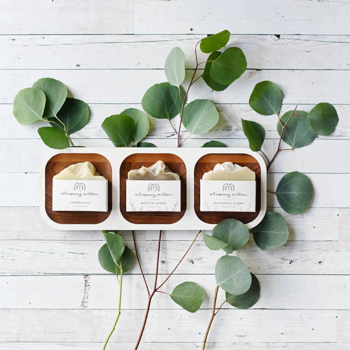 Three small soap bars on a wooden tray with eucalyptus leaves on a white wooden surface
