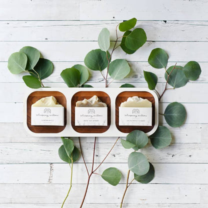 Three small soap bars on a wooden tray with eucalyptus leaves on a white wooden surface