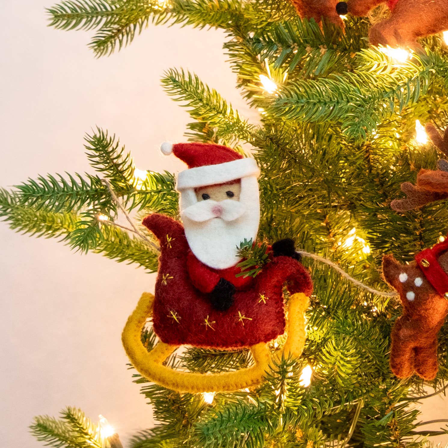 A felt Santa Claus sitting in a sleigh, hanging on a lit green Christmas tree. The ornament features red clothing, a yellow sleigh, and small gold star details.
