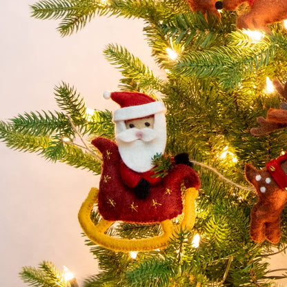 A felt Santa Claus sitting in a sleigh, hanging on a lit green Christmas tree. The ornament features red clothing, a yellow sleigh, and small gold star details.