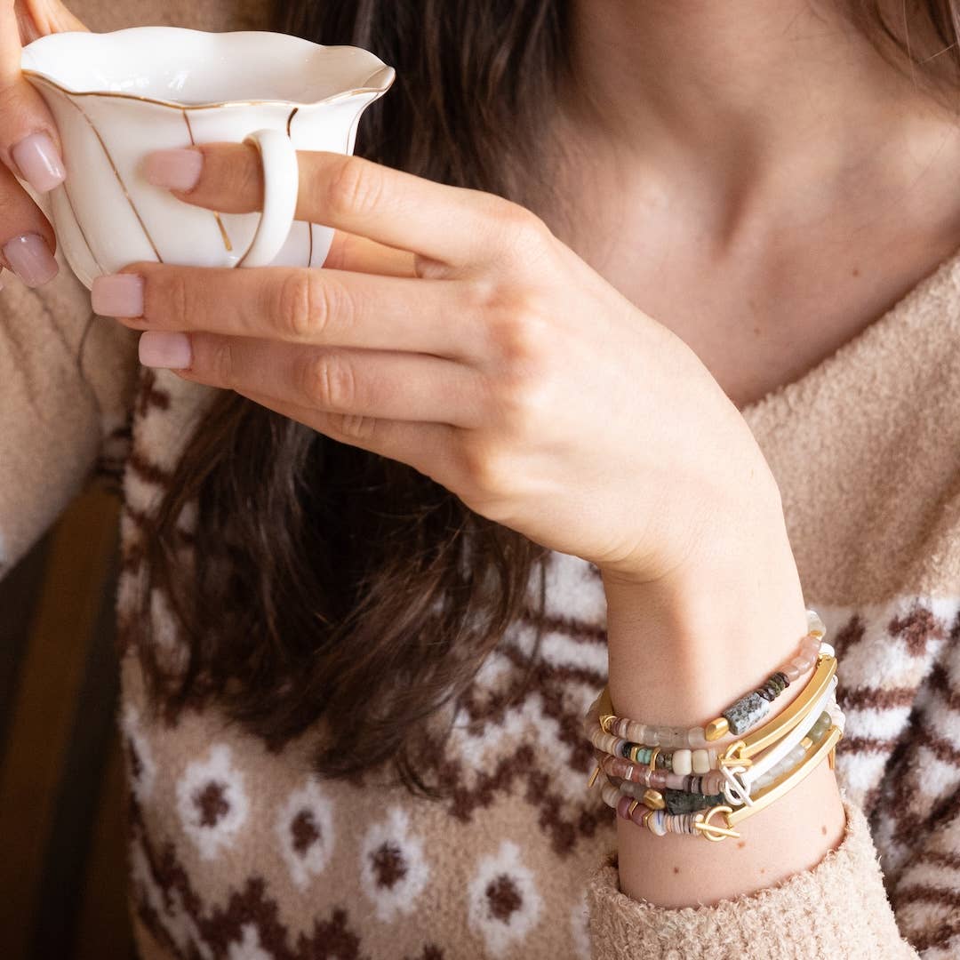 Person holding a small white ceramic cup with a patterned sweater and bracelets.