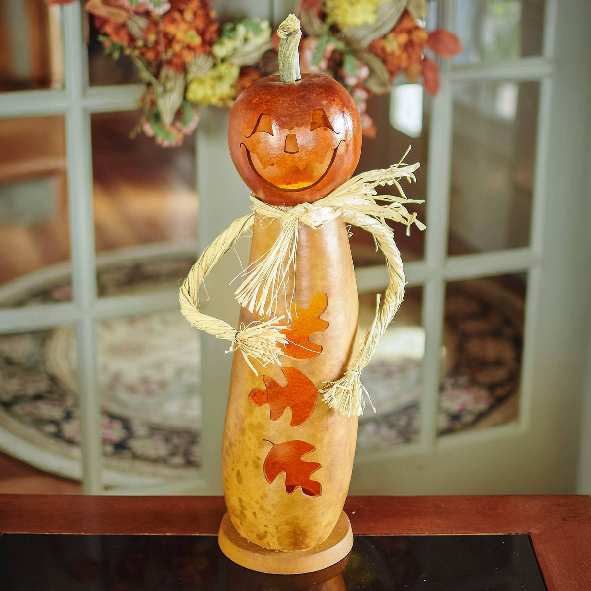 A decorative carved gourd figure with a pumpkin head, straw-colored stem and scarf, and orange leaf cutouts on the body, displayed indoors on a table in front of a window.