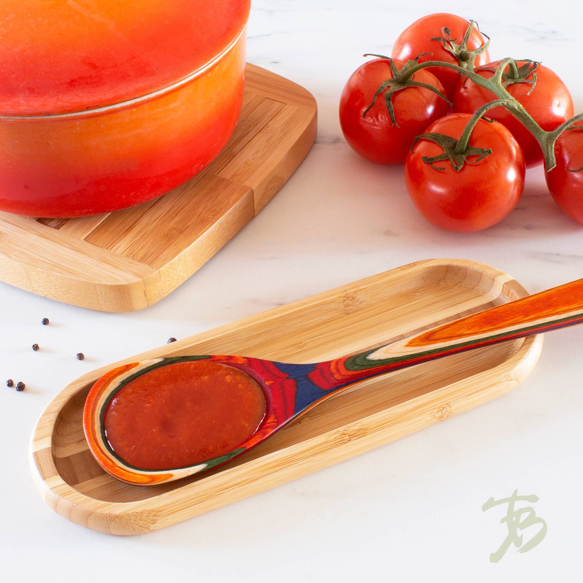 Wooden spoon with red sauce on a wooden tray, surrounded by tomatoes and a pot.