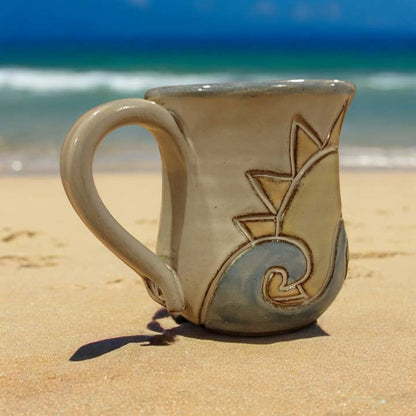 Ceramic mug with a sun and wave design on a sandy beach with ocean in the background