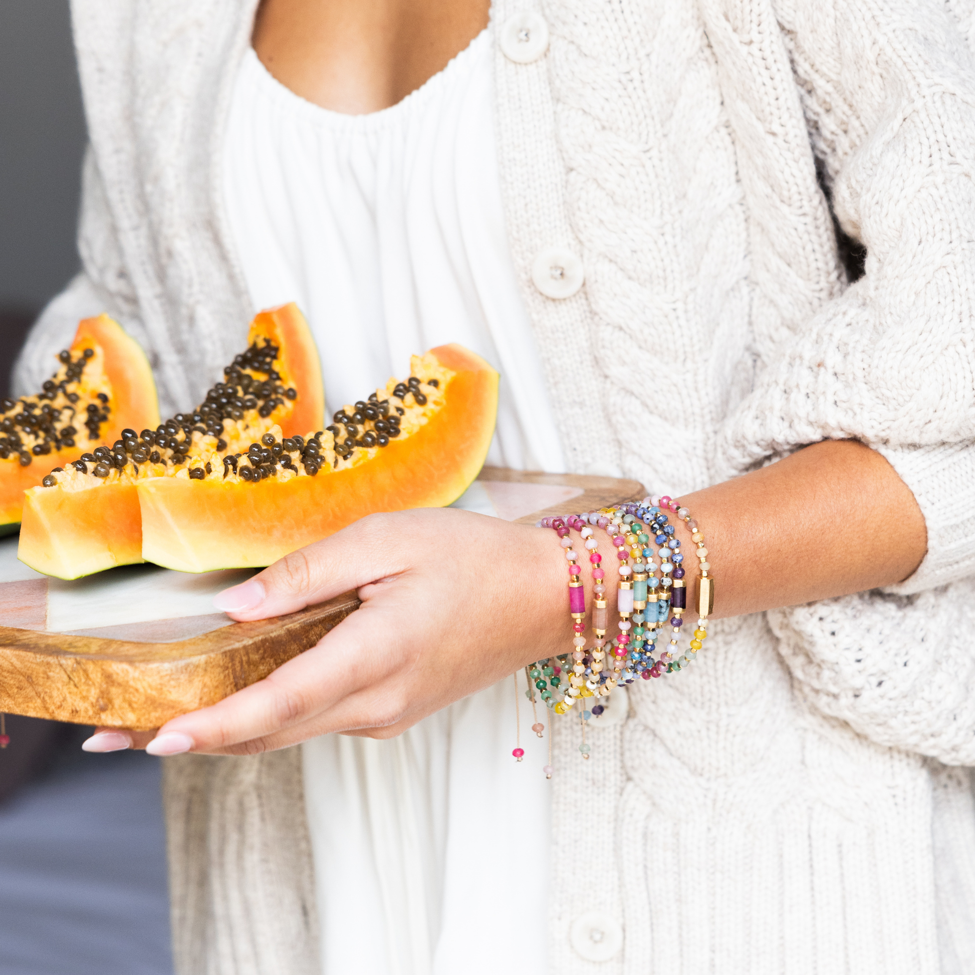 Person holding a wooden plate with papaya slices and colorful bracelets on a neutral background