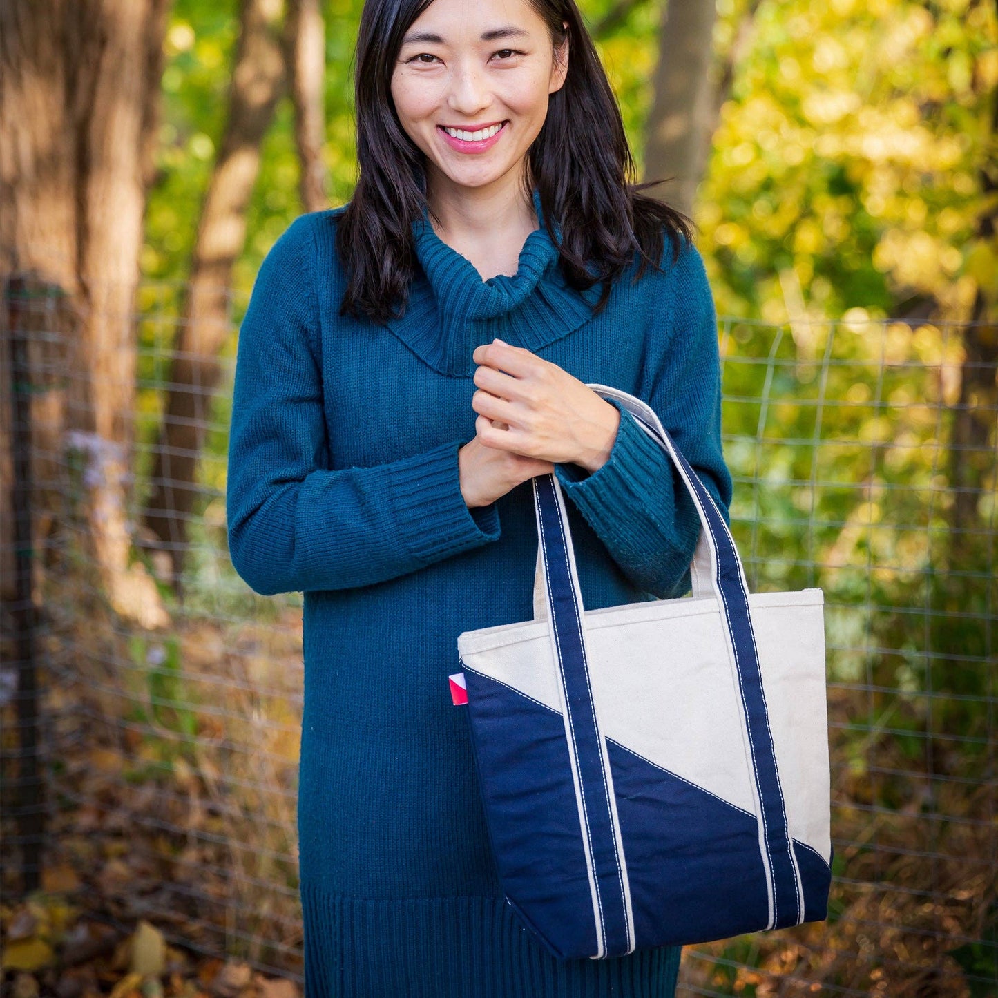 Woman holding a blue and white tote bag outdoors with greenery in the background