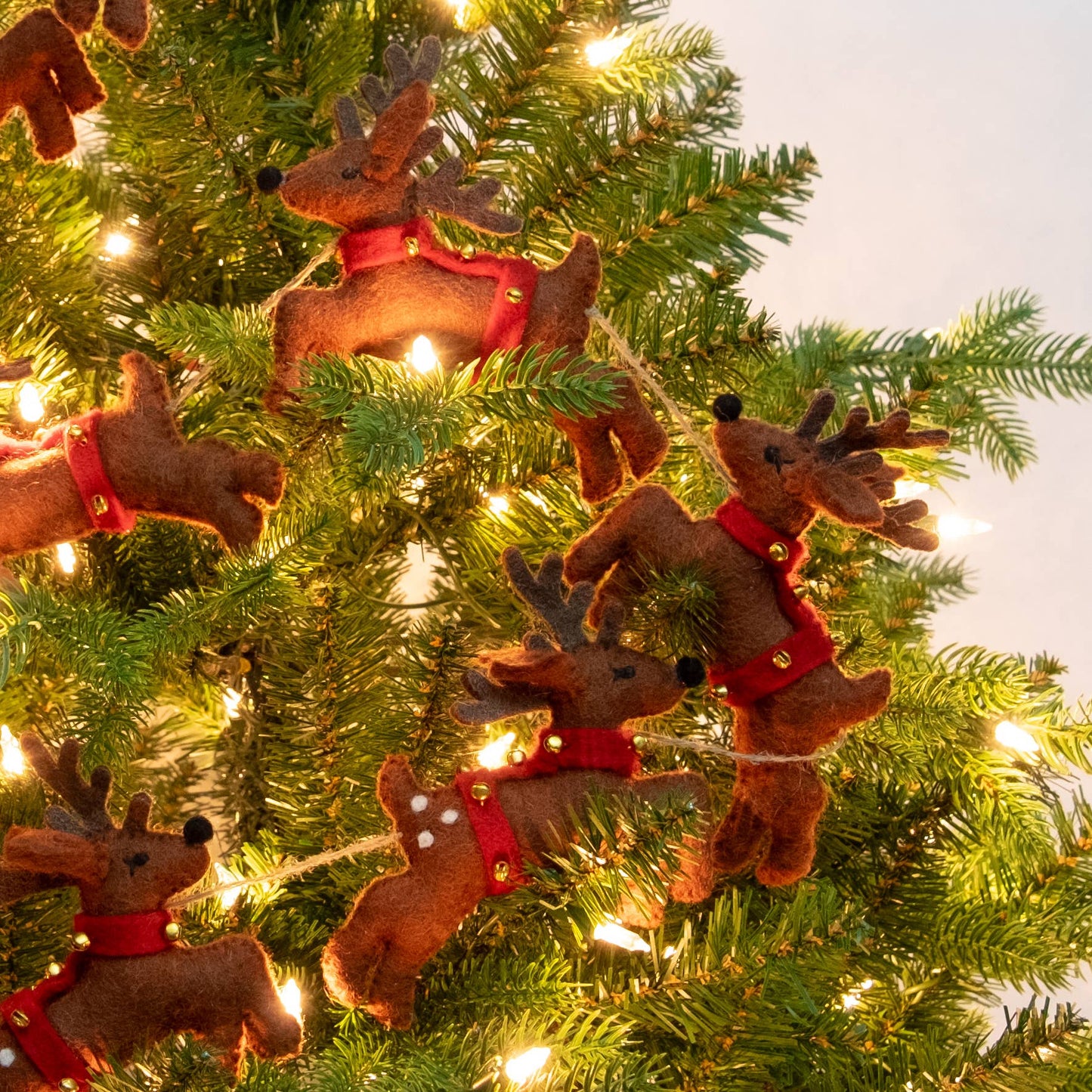 Felted brown reindeer Christmas garland with red harnesses and gold bells hanging on a green Christmas tree with warm white lights.