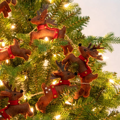 Felted brown reindeer Christmas garland with red harnesses and gold bells hanging on a green Christmas tree with warm white lights.