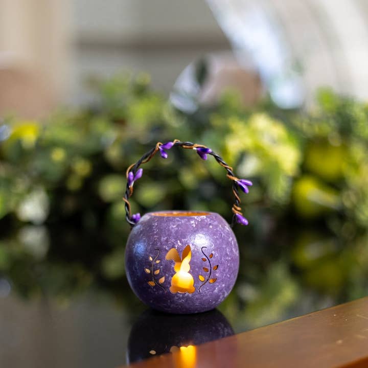 Purple decorative lantern with a lit candle inside on a reflective surface with a blurred green background
