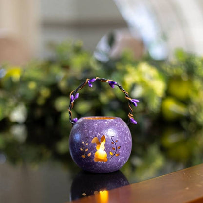 Purple decorative lantern with a lit candle inside on a reflective surface with a blurred green background