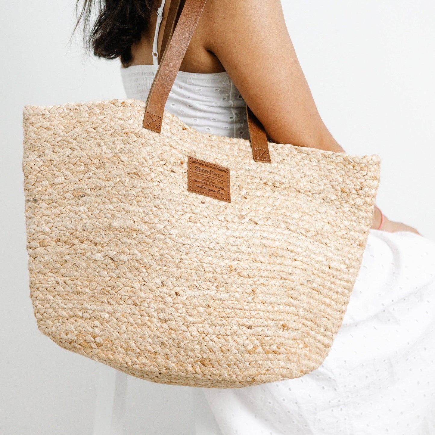 Woven straw tote bag with brown leather handle and patch held by a person against a white background