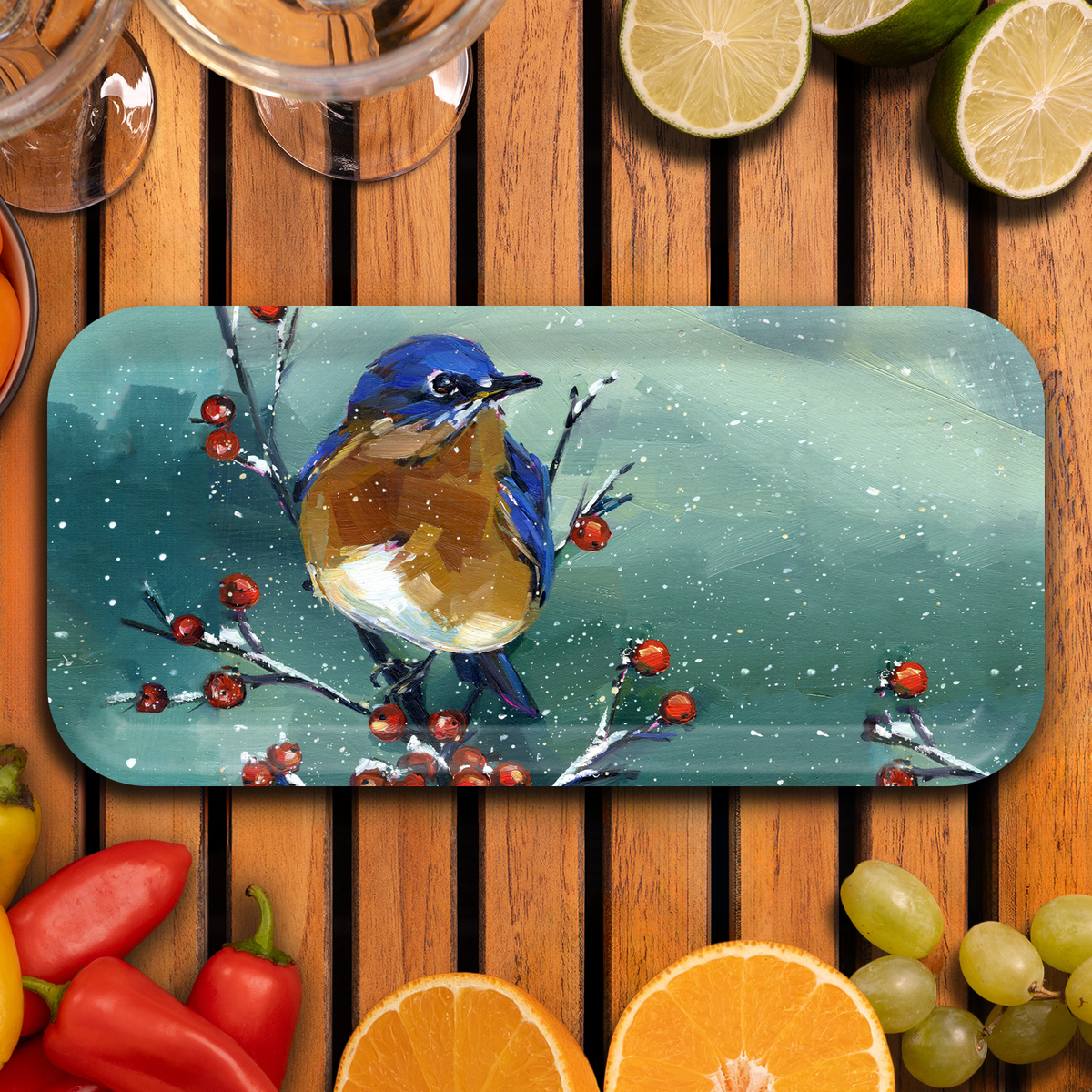 A rectangular tray with rounded corners featuring a painted bluebird scene, displayed on a wooden table surrounded by sliced limes, grapes, peppers, and citrus.