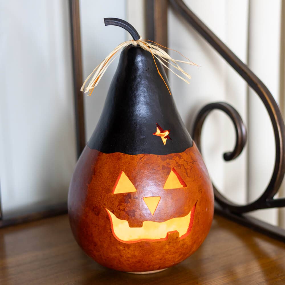 A carved orange pumpkin gourd with a black pointed hat and a small star detail, lit from within to glow with warm light, set on a wooden surface.