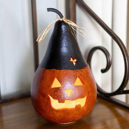 A carved orange pumpkin gourd with a black pointed hat and a small star detail, lit from within to glow with warm light, set on a wooden surface.