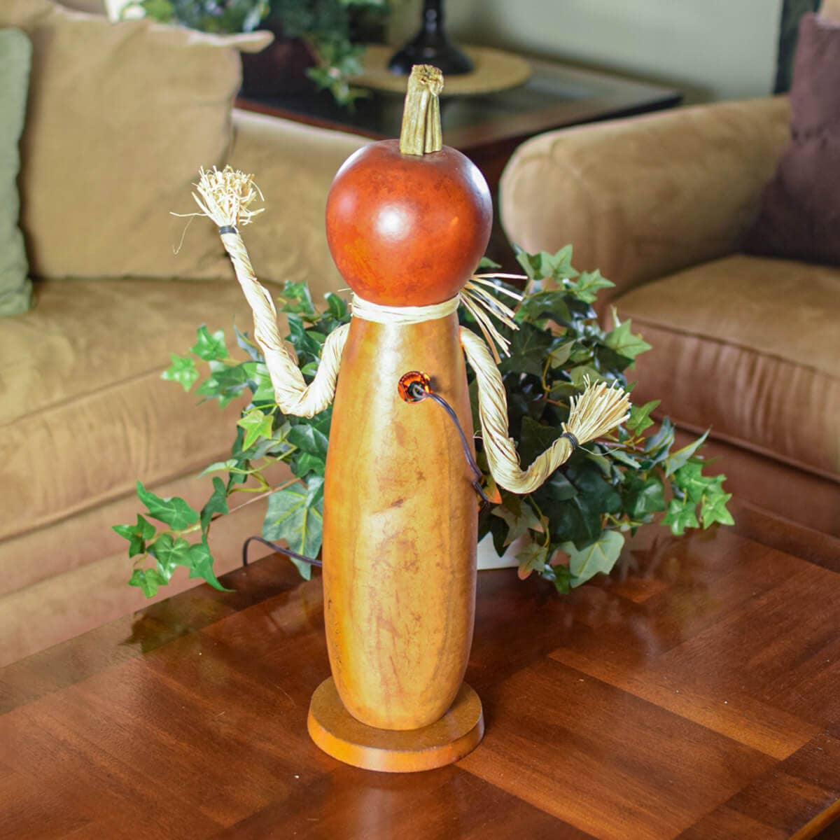 A handmade wooden and gourd scarecrow decoration displayed on a wooden table in a living room setting, with a brown leather sofa and green ivy plants in the background.